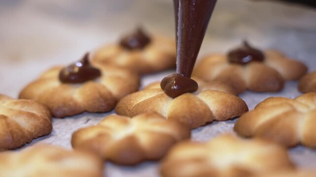 A baker uses a piping bag to add chocolate icing to flower-shaped cookies on parchment paper, showcasing the meticulous decoration process in a well-lit kitchen environment