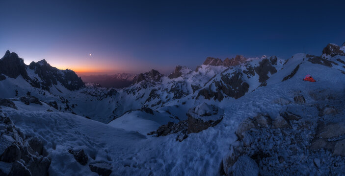 View of a snow-covered mountain range at twilight with a small orange tent pitched on a snowy slope in Spain.