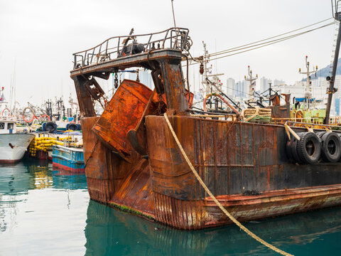 View of a weathered, rusty fishing boat moored in the harbor with other vessels and the urban skyline in the background in Busan, Busan, South Korea.