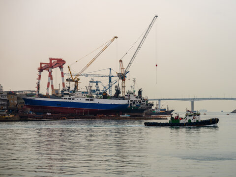 View of a shipyard with large cranes, a ship under construction, and a tugboat navigating the harbor in Busan, Busan, South Korea.