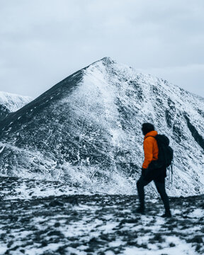 View of Catstycam peak covered in snow with a hiker in an orange jacket walking along the ridge in the Lake District, Penrith, England, United Kingdom.