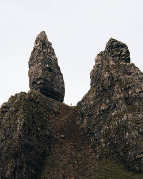 View of The Storr, a dramatic rocky pinnacle with a small person standing on the slope below under an overcast sky in Portree, Scotland, United Kingdom.