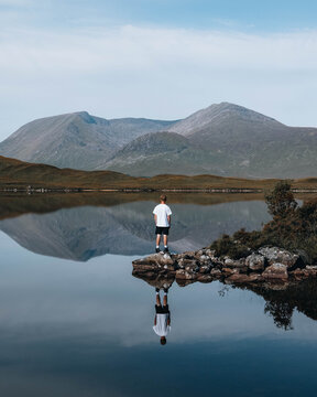View of a man standing on a rock in a calm lake with a clear reflection and the Scottish Highlands in the background in Fort Augustus, Scotland, United Kingdom.