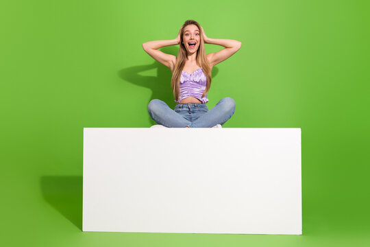Happy young glamorous female model sits cross legged on a white board against a green background in casual fashion style