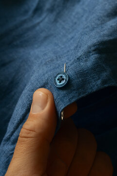 Detailed close-up of a hand sewing a blue button onto blue linen shirt fabric with needle and thread, tailor crafting process shot, organic textile background, sustainable fashion repair, mending.