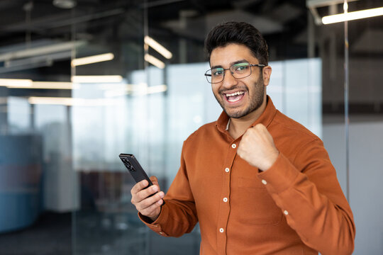 Successful businessman celebrating achievement with smartphone in office, happy young man looking at phone screen with excitement and winning gesture for business growth.