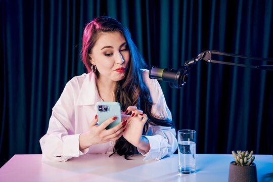 Female content creator recording a podcast while checking smartphone in studio with microphone and glass of water, lifestyle streaming and social media concept