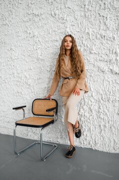 Beige blazer wearing woman leaning against rugged textured white wall beside vintage chair, fashion forward pose showcasing neutral palette and contemporary street style