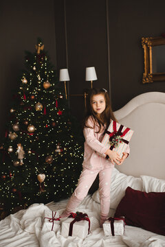 Smiling child girl 5-6 year old holding Christmas present box in bed over xmas tree in bedroom. Winter holiday season.