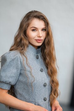 Portrait of woman with long wavy brown hair wearing gray wool coat with decorative metal buttons, soft neutral backdrop, elegant pose, natural makeup, studio lighting, close crop