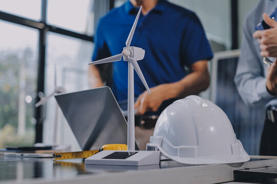 Two Asian engineers wearing hardhats and safety vests discussing solar panel installation and renewable energy project in the office.