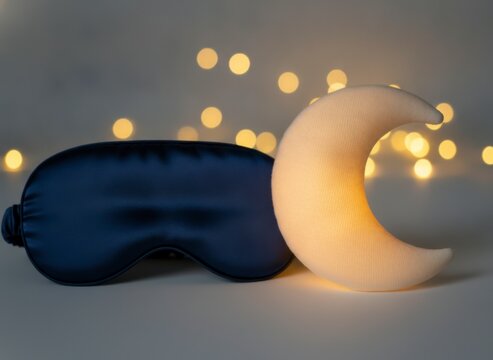 Blue silk sleep mask next to a glowing crescent moon night light with warm bokeh lights in the background, symbolizing restful sleep