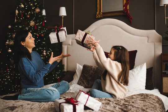 Happy woman sitting in bed with kid girl open Christmas present boxes in bedroom. Winter holiday season. Cozy home atmosphere.