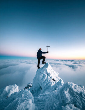 A determined mountaineer stands atop a snowcovered peak holding an ice axe above a sea of clouds at sunset. This inspiring image symbolizes ultimate success, peak performance, and personal growth.