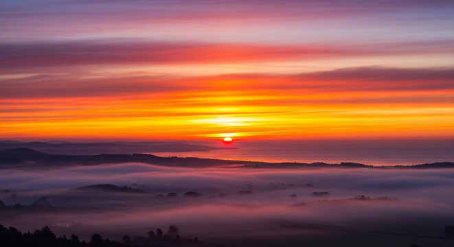 A vibrant sunrise paints the morning sky with brilliant shades of orange and purple while a thick layer of low fog blankets the rolling hills below during a peaceful early dawn hour