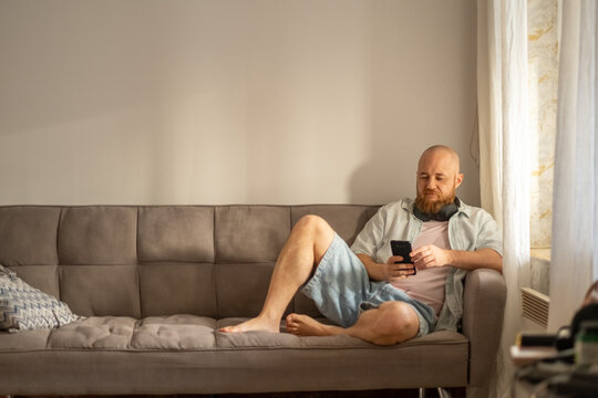 Bearded man sits alone with phone in warm sunlit living room, leisure spent with screen instead of book, quiet hour worth protecting. Wide shot. Concept of leisure evening of adult man