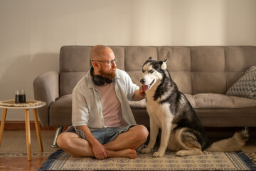 Man sits cross legged on carpet stroking husky next to grey sofa, affection that both of them need, warmth of shared home morning. Wide shot. Concept of daily affection between owner and dog © iwavephoto