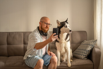 Man extends arm taking selfie with husky sitting beside him on sofa, blogging about daily life with pet, comfort of living with dog. Wide shot. Concept of blogging with dog for social media © iwavephoto
