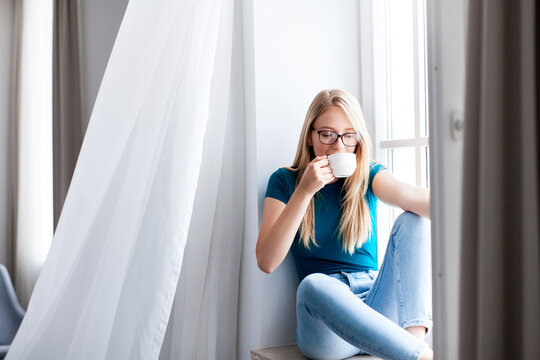 Young woman drinking coffee at home, relaxing on windowsill. Girl breathing fresh air, enjoying calming beverage, morning wind from opened window. Recovery, mental health, slow living, mindfulness