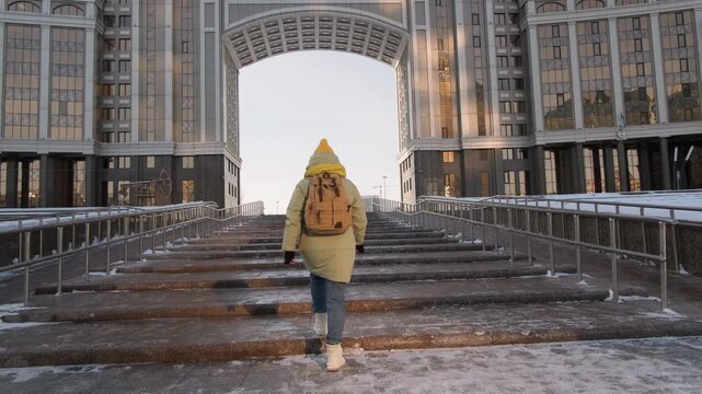 Woman tourist climbs street staircase against background of arched passage in building while exploring Astana in winter