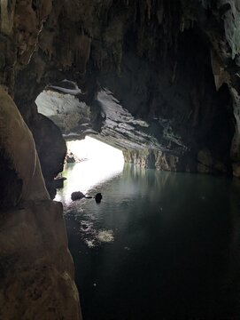 Looking from the cavern darkness toward a brightly lit cave exit over calm subterranean waters reflecting daylight.