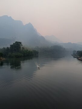 Atmospheric landscape view of the Nam Ou river flanked by tall jungle-covered limestone karst mountains under a hazy sky in Nong Khiaw, Laos.