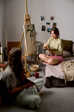 Vertical shot of young adult Caucasian couple engaging in creative hobbies, woman painting while partner playing guitar. Concept representing home lifestyle, ideal for youth culture marketing