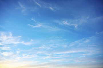 Fototapeta premium Light white cirrus clouds scatter across bright blue sky with soft yellow gradient near horizon. Clear atmospheric texture background