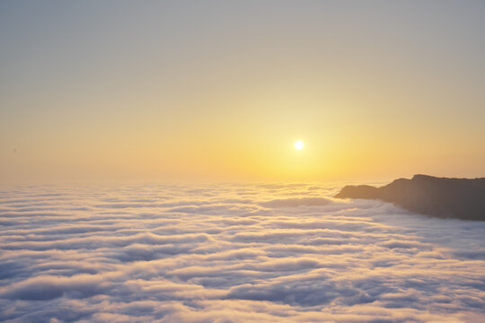 Bright sunrise sun shines over dense rolling clouds layer near dark mountain peak rocky silhouette. Golden dawn landscape