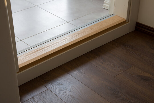 Light wooden threshold installed at a doorway to connect two different flooring materials. The transition smoothly bridges the gap between dark wood laminate planks and light grey ceramic floor tiles.