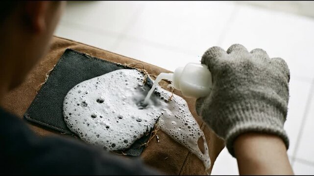 Gloved hand dispensing white foamy liquid onto a dark denim patch on brown fabric in bright indoor light
