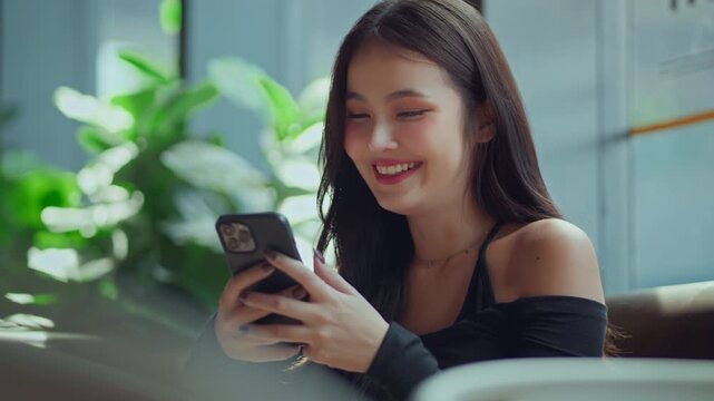 Happy young Asian woman smiling while using smartphone at home, cheerful female reading message and browsing mobile app, online communication, social media.