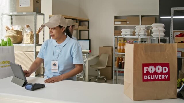 Young Hispanic woman managing orders on digital tablet with Caucasian man packing groceries into delivery bag. Perfect for food delivery and e-commerce advertising