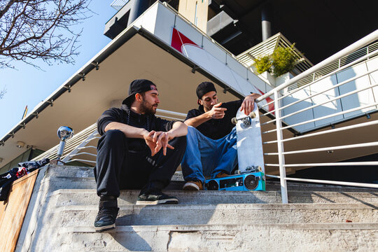 Two young male skateboarders enjoying conversation while sitting on concrete stairs in the city