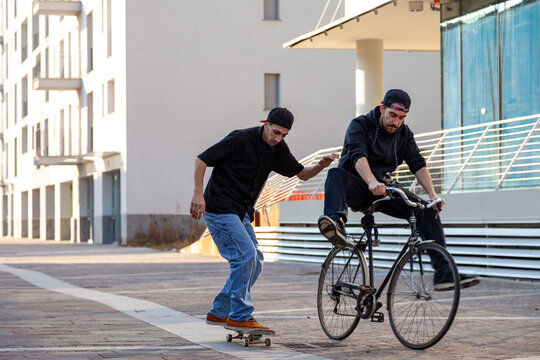 Two young men performing tricks on a skateboard and bicycle outdoors in a city setting