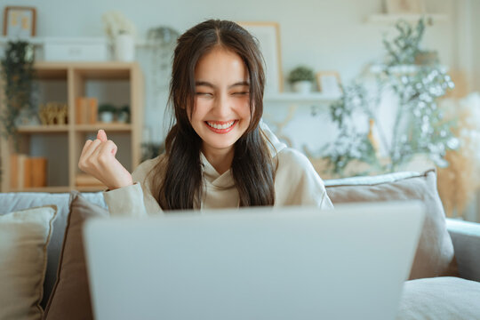 Excited young woman celebrating success while using laptop on sofa at home, smiling with happiness after positive online result, achievement, good news.
