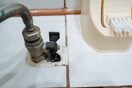 Aged bathroom water shutoff valve closeup beside plastic brush holder on tiled floor, showing metal flexible hose, grout lines, stains, and worn plumbing hardware