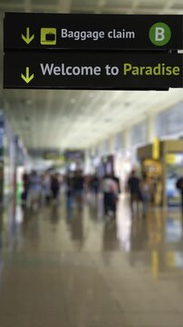 Close up view of an airport arrival sign with baggage claim directions and a Welcome to Paradise message, with a blurred background of passengers walking through the terminal hall