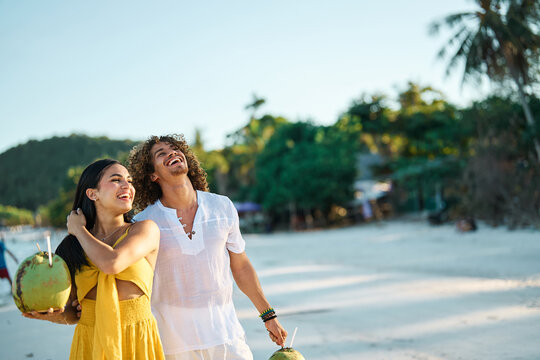 A smiling couple strolls barefoot along a tropical beach, each holding a fresh coconut as gentle waves lap the shore. The woman wears a yellow dress and the man white linen clothing, creating a relaxe