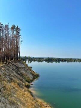 Steep Sandy Cliff with Pine Trees Above Turquoise Quarry Lake Vertical