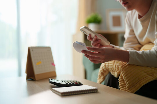 Woman comparing bills with information on a smartphone beside a calculator and calendar at home, personal debt, monthly payments, repayment deadlines, household expenses, budgeting financial pressure