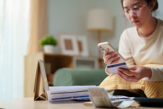 Woman checking a credit card with smartphone while reviewing bills and money at home, personal debt, credit card payment, household expenses, online banking, financial stress and debt management