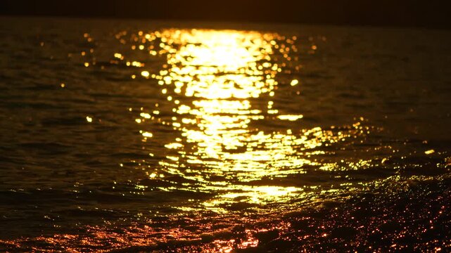 Shimmering sunset path across dark ocean water. Static close up shot captures bright golden light reflecting on rippling sea surface.