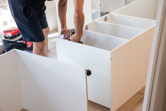 Worker assembling white kitchen cabinet carcass on floor, modular furniture construction and home renovation.