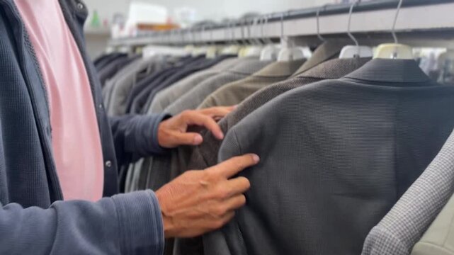 Close-up view of a man browsing through a rack of secondhand suits and jackets while shopping for affordable clothing at a thrift store. 15416  