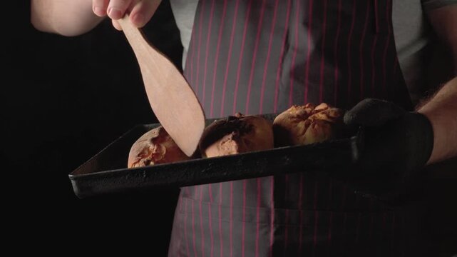 Male baker in striped apron uses wooden spatula to glaze freshly baked bread rolls on a tray, showcasing the intricate details of the bread and the baking process in a dark kitchen setting