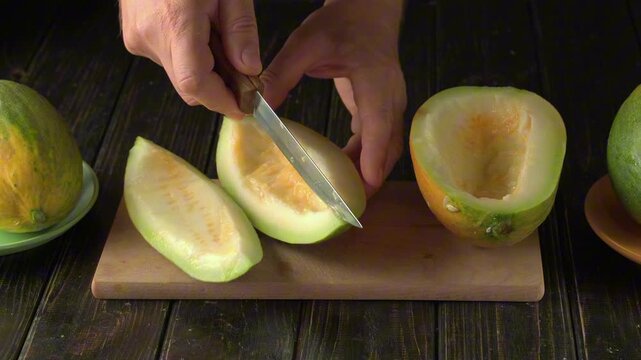 Hands slicing a green melon on a wooden cutting board, revealing its inner flesh while additional melons rest on the table, showcasing the process of preparing fresh fruit for consumption