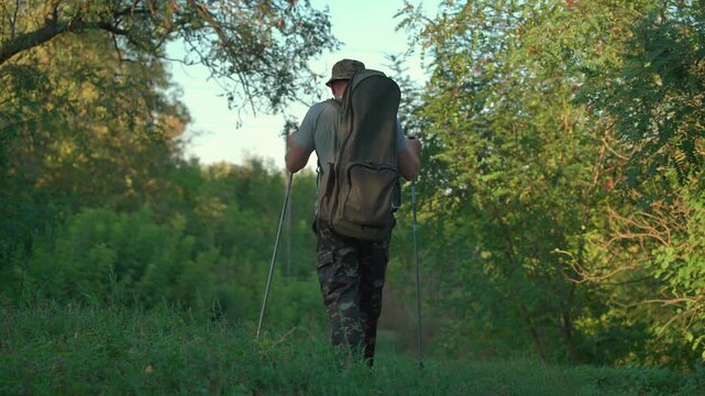 Male hiker with camouflage pants and backpack uses trekking poles while walking through a lush green forest path surrounded by trees and underbrush in a natural outdoor setting