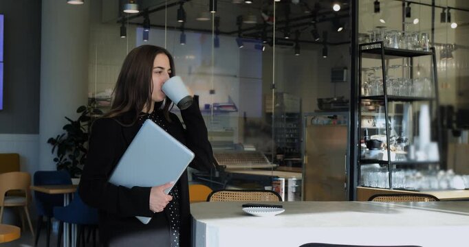 Exhausted woman holding laptop sips hot beverage in contemporary coffeehouse. Overworked corporate employee pauses beside white table feeling work burnout after long hours of business tasks.