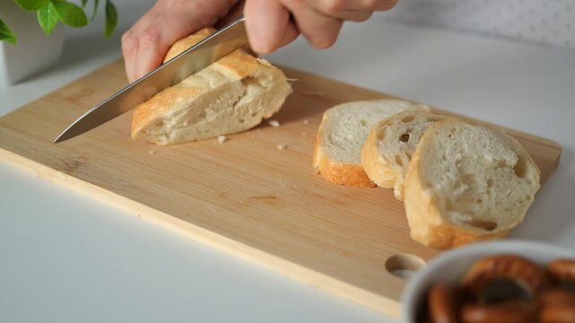 Slicing freshly baked baguette loaf with stainless knife on cutting board aids wholesome breakfast preparation. Skilled hands cut bread slices steadily while preparing delicious morning food.